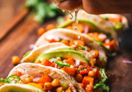 A hand squeezing lime over a row of tacos filled with chickpeas, diced vegetables, and avocado; garnished with cilantro on a wooden board.