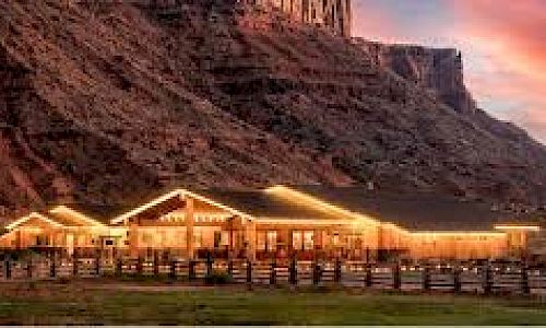 A warmly lit house is set against a mountainous backdrop, with a sunset sky and a wooden fence in the foreground.